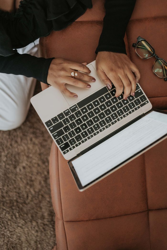 services-01 A woman wearing a ring types on a laptop on a brown couch with eyeglasses beside her.