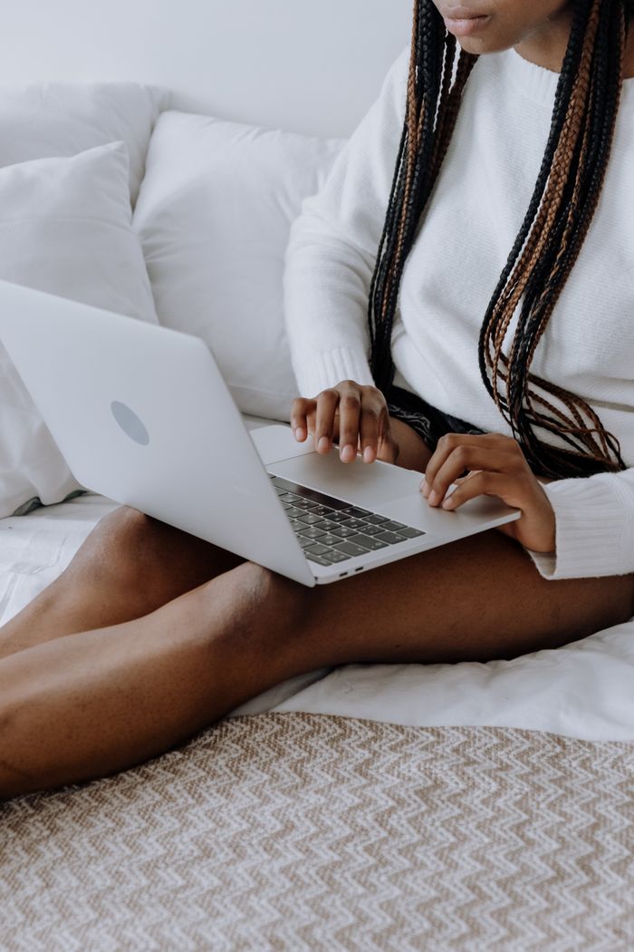 about-img Young woman typing on laptop while sitting on bed in a cozy home setting. Ideal for technology and lifestyle themes.
